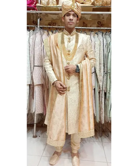 A full-length view of a groom in a light beige silk sherwani. The outfit is accessorized with a gold mala, showcasing a complete and elegant look available for rent.