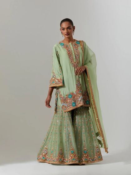 A full-length view of a model wearing a pastel green sharara set. The kurta is heavily embroidered with floral motifs in shades of orange, blue, and gold.