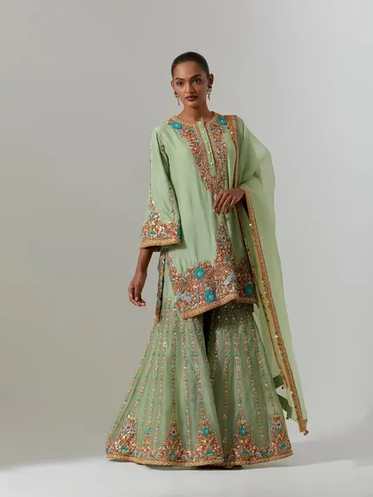 A full-length view of a model wearing a pastel green sharara set. The kurta is heavily embroidered with floral motifs in shades of orange, blue, and gold.