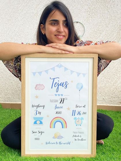 I love holding the finished product. Here I am with a framed baby biography for Tejas, featuring a blue color scheme with a balloon and rainbow. It’s so rewarding to create these special keepsakes for new parents.