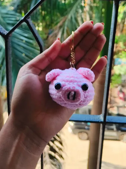 Holding the little pig keychain in my hand. You can see the texture of the crochet stitches and the secure gold-colored keyring I use.