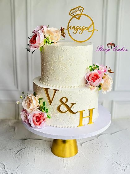 An elegant two-tier engagement cake with a subtle floral texture. The design is personalized with the couple's initials in gold and adorned with beautiful bouquets of pink and white roses.