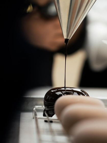 A close-up of a dessert being glazed with dark chocolate, capturing the smooth, liquid texture of the glaze.