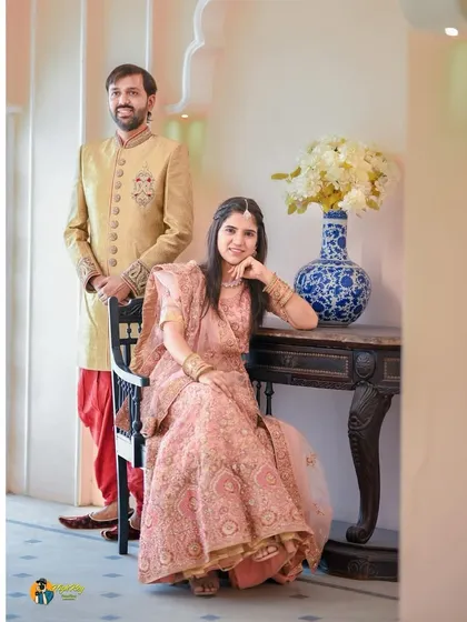 A regal portrait of a couple in traditional Indian attire, posing in a room with ornate, palace-like decor. The groom stands in a classic sherwani while the bride is seated, creating a timeless and elegant composition.