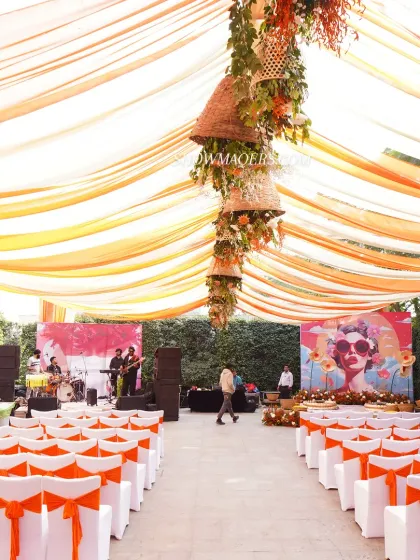 We love creating vibrant, high-energy Haldi ceremonies like this 'Art of Orange' themed event. The setup features a canopy of orange and yellow drapes, a quirky pop-art style backdrop, and unique floral installations hanging from wicker baskets, all designed to create a fun and festive atmosphere.
