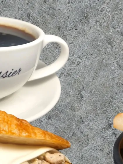 Another view of a perfect pairing. A cup of black coffee from a branded mug sits next to a delicious looking croissant sandwich, ready to be enjoyed.