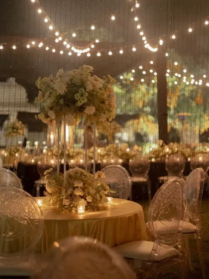 A moment that feels like poetry. This intimate dinner setting combines the golden glow of string lights and candles with ethereal white floral centerpieces and modern ghost chairs for a magical feel.