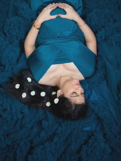 An artistic top-down shot of a pregnant woman lying on the train of her blue gown, with flowers in her hair.