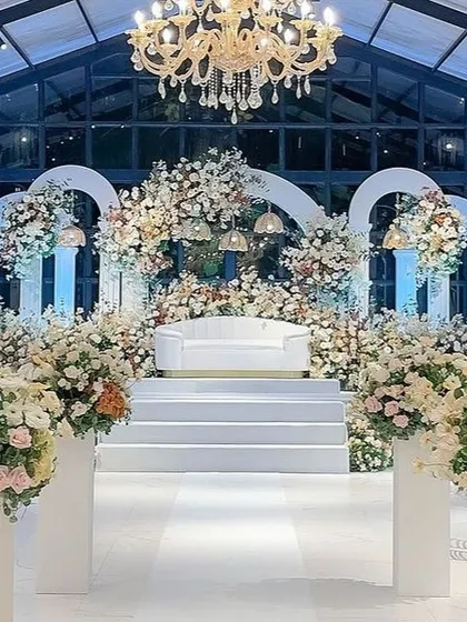 Set within a glasshouse, this all-white stage combines classic architectural arches with an abundance of flowers and elegant lighting. The design feels both grand and intimate, making it a perfect setting for a sophisticated wedding reception.