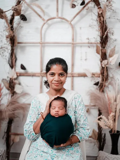 A mother proudly presents her newborn to the world. She holds her peacefully sleeping baby, both looking towards the camera, in a classic and heartfelt portrait.