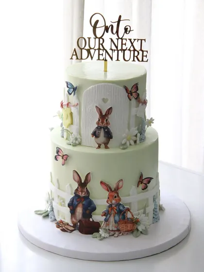 "Onto our next adventure." This charming two-tiered Peter Rabbit themed cake was created for a baby shower, celebrating the beautiful journey of welcoming a new family member.