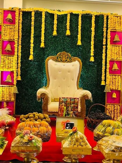 A vibrant Seemantham stage with a checkerboard backdrop in yellow and magenta. The decor is enhanced with hanging marigold garlands and brass bells, with a full display of offerings in the front.
