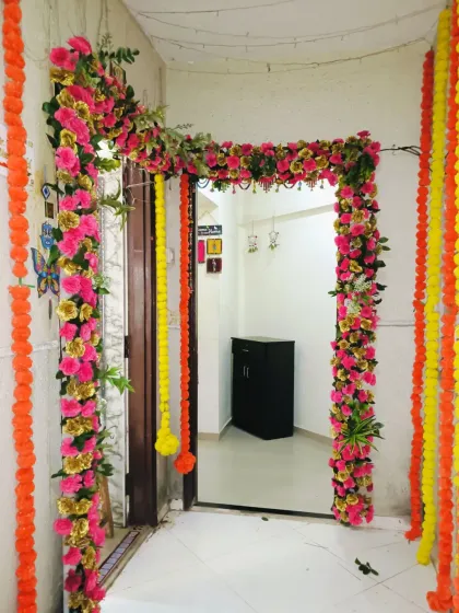 A festive floral decoration for a home entrance, perfect for a housewarming, engagement, or any pooja. The doorway is framed with pink and yellow artificial flowers, complemented by hanging marigold garlands.