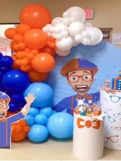 An energetic Blippi themed birthday party with his signature orange and blue colors. The setup includes a large character cutout, a themed backdrop, and custom pedestals decorated with glasses and bowties.