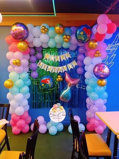 A mermaid-themed birthday party setup in a restaurant, with a colorful balloon arch and shimmer backdrop.