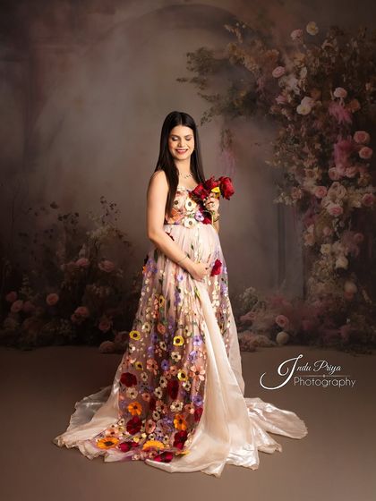 A gentle and serene portrait of a mom-to-be in a gown adorned with colorful, three-dimensional flowers. The soft lighting and floral backdrop create a romantic, painterly feel.