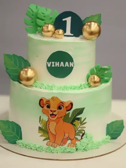 A two-tier Simba themed cake for a "Wild One" first birthday. The light green frosting and jungle leaves create a perfect backdrop for the little lion king.