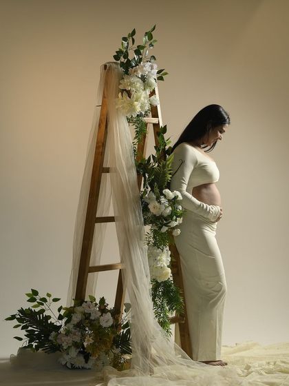 A portrait of quiet strength. The mother-to-be stands beside a ladder adorned with white flowers, her profile illuminated by soft studio light, creating a serene and symbolic image of growth.