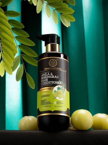 A dark and moody shot of a Khadi Natural hair conditioner. The rich green background and fresh amla fruits highlight the product's natural, ayurvedic ingredients.