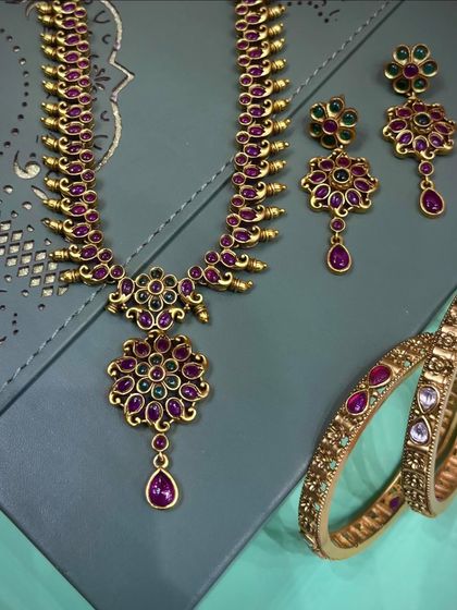 This antique-finish necklace set features intricate floral motifs with ruby and emerald colored stones. The matching earrings and bangles complete the traditional look.