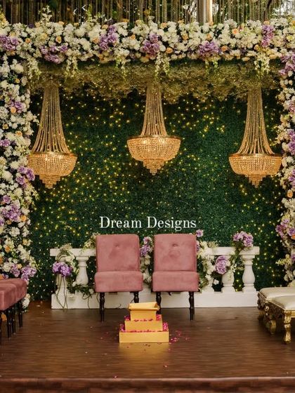 This mandap combines a lush green backdrop with a frame of purple and white flowers. The three hanging crystal chandeliers and traditional golden pillars add a regal touch to the sacred space.