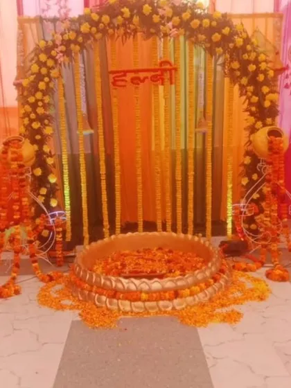 A traditional Haldi ceremony setup with a floral arch and a large brass urli. The decor uses marigold garlands and decorative matkas on stands, perfect for a home-based function.
