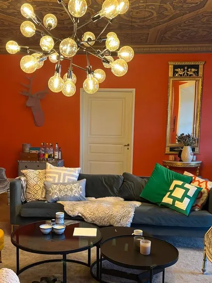 A vibrant living room where my custom table runner, carpets, and cushion covers fit perfectly. The bold orange walls are balanced by the deep green sofa and the eclectic mix of my handcrafted textiles.