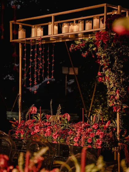 A modern and elegant decor element from the red Sangeet. A gold metal structure holds candles above a table laden with red flowers, with delicate red floral strings hanging down.