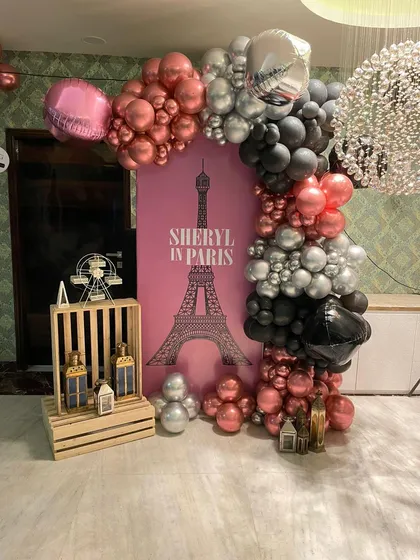 A stylish "Sheryl in Paris" themed photo corner with a pink backdrop, an Eiffel Tower graphic, and a rose gold, silver, and black balloon arch.