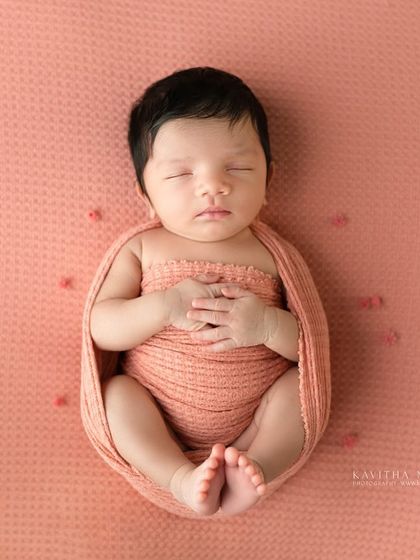 A one-week-old baby girl wrapped in a peachy-pink swaddle. The simple, clean background allows the baby's peaceful expression to be the main focus.
