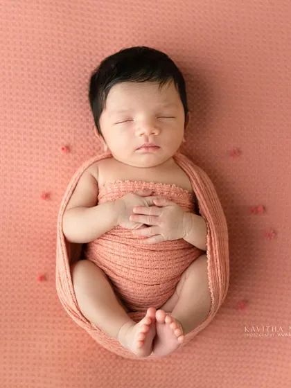 A one-week-old baby girl wrapped in a peachy-pink swaddle. The simple, clean background allows the baby's peaceful expression to be the main focus.