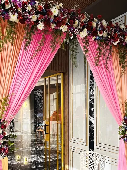 The entrance decor for the Godhana and Chunadi ceremony, with bright pink drapes and colorful floral bouquets creating a festive and welcoming entryway.