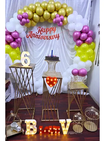 I design romantic decorations for anniversaries. This setup features a beautiful balloon arch, elegant gold-toned stands for the cake, and marquee letters to celebrate the special occasion.