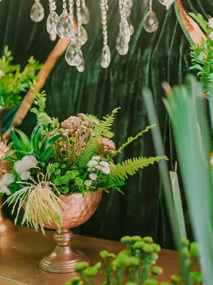 A close-up of a floral arrangement featuring a mix of ferns, white flowers, and other greenery in a copper vase, set against the rich texture of a velvet drape.