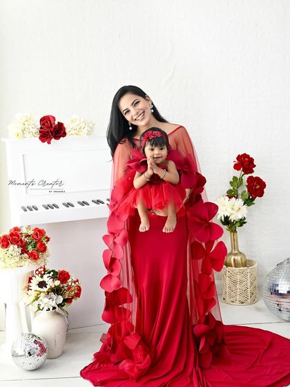 A beautiful mother-daughter moment in matching red dresses. The little one's "namaste" gesture is incredibly sweet, captured against a clean, bright studio setup with a white piano.