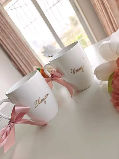 A pair of ceramic mugs personalized with the couple's names in gold calligraphy. A simple yet elegant anniversary gift, tied with a soft pink ribbon.