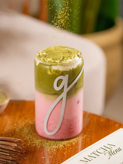 A layered strawberry matcha latte, with matcha powder being sprinkled on top. The action adds a finishing touch.