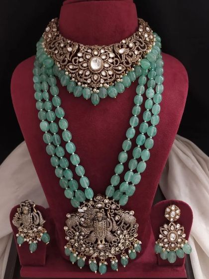 This pastel green jewelry set features a choker with a large kundan-style centerpiece and a long beaded necklace with a deity pendant. It's a beautiful mix of different traditional styles.