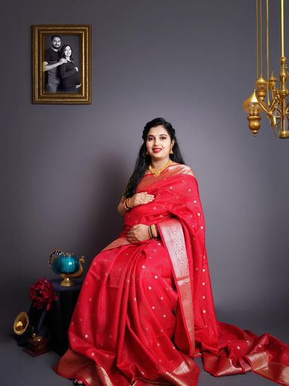 A classic studio portrait of a mother-to-be in a red silk saree, with a framed photo of her and her husband in the background.