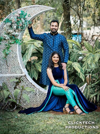 A romantic pose with a couple by a large crescent moon prop in a garden, dressed in elegant blue traditional outfits.