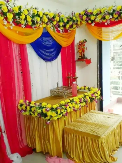 A simple and colorful Ganpati pooja corner for a home. This setup uses bright fabric drapes in yellow, pink, and blue, with yellow floral arrangements on the table and as a top border.