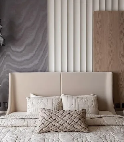 A masterclass in modern tranquility, this bedroom uses a layered palette of textures to create a sensory experience. The subtle wave pattern of the wallcovering, paired with a clean-lined upholstered headboard and woven cushions, exudes a sense of stillness and calm.