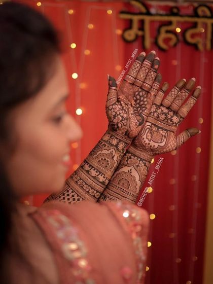 The bride proudly displays her full bridal mehendi, which features beautiful peacock and elephant motifs, in front of a festive "Mehndi" backdrop.