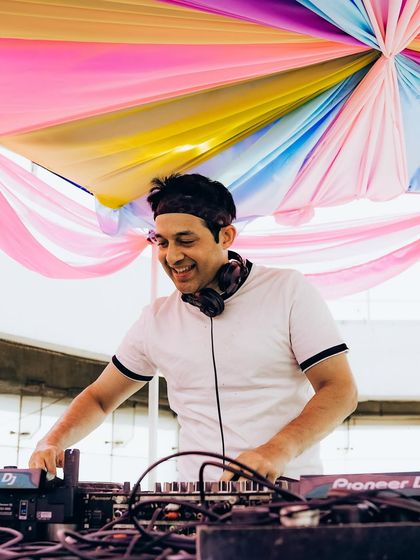 A DJ smiles while spinning tunes under a colorful canopy at our rooftop Holi party, capturing the joyful and vibrant atmosphere we create.