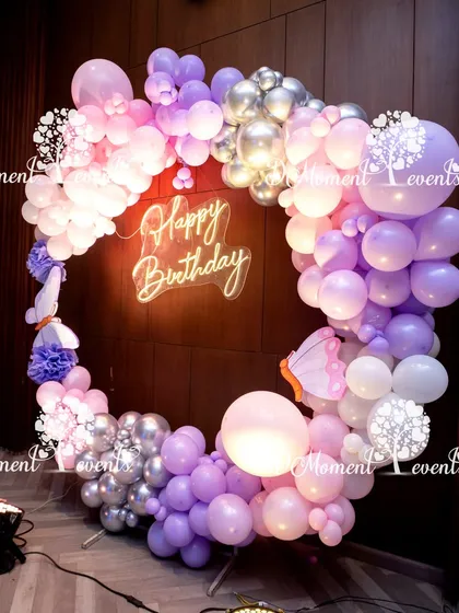 A heart-shaped balloon arch in shades of pink, purple, and silver. The "Happy Birthday" neon sign in the center makes this a perfect, modern photo booth.