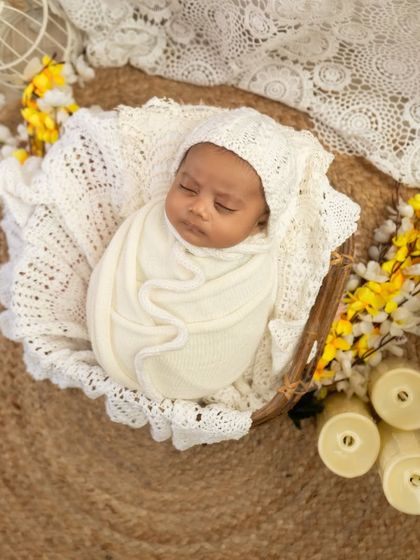 A peaceful portrait of a baby in a white bonnet and wrap, resting in a basket on a jute mat with lace and floral details.