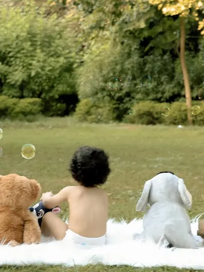 Photographed from behind, a baby sits with his teddy bear friends, watching bubbles float by. This image tells a story of friendship and wonder, perfect for a first birthday theme.