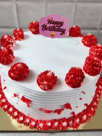 A classic red velvet birthday cake with simple and cheerful decorations. The cake is frosted in white with red crumb accents and piped red rosettes, topped with a "Happy Birthday" sign.