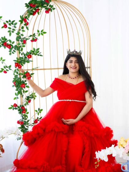 A whimsical portrait in our studio. The mom-to-be in a red gown sits next to our golden birdcage prop, adorned with climbing red roses, creating a magical, storybook feel.