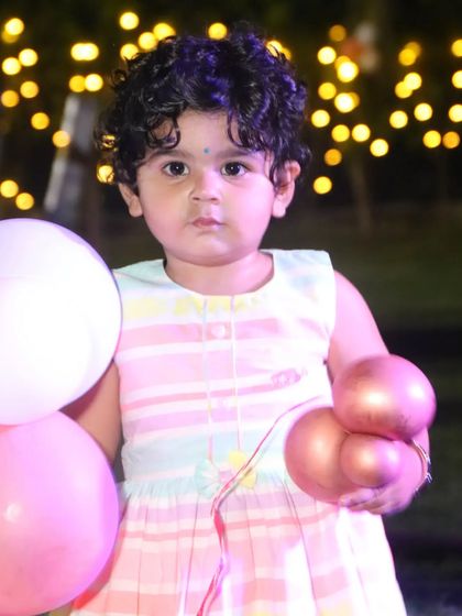 A little girl in a striped dress holds balloons, captured against a soft-focus background of party lights.